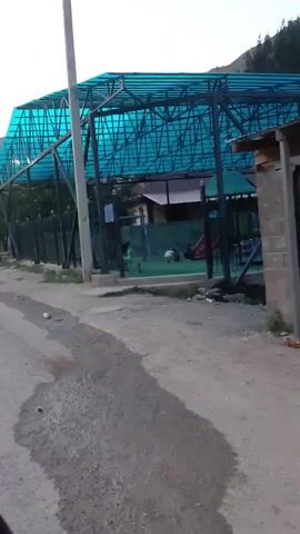 This video captures a serene, rural setting in Terek-Say, Kyrgyzstan, likely during the late afternoon or early evening as indicated by the soft, diffused light and the presence of streetlights which are beginning to illuminate. The scene is dominated by a dusty, unpaved road that winds through a village nestled at the foot of imposing mountains.

Prominently featured is a modern, covered sports court or recreational area with a vibrant turquoise, translucent roof supported by a dark metal frame. Adjacent to this structure, a children's playground with a red slide is visible, suggesting a community space. The road itself shows signs of recent moisture, with dark, wet patches indicating puddles or dampness.

The surrounding environment is characterized by modest, rustic buildings, some appearing to be agricultural structures with wooden supports and corrugated metal roofing. The natural landscape is a key element, with the rugged, barren mountains forming a dramatic backdrop to the village.

There are a few individuals visible, including a person walking in the distance on the left side of the road and some figures in the playground area. The overall mood is peaceful and quiet, conveying a sense of everyday life in a remote, mountainous region. The lack of dense population and the simplicity of the structures suggest a close-knit community. The weather appears to be clear, with a hazy sky that is typical of the fading daylight. No significant text is visible within the frames.