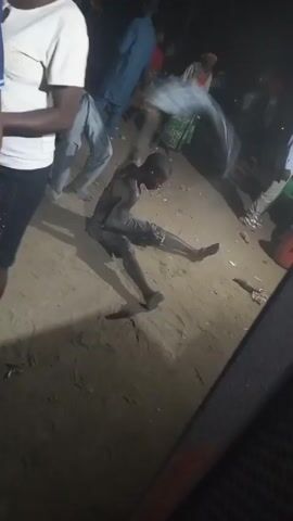 The video depicts a man sitting on the ground in an outdoor setting, surrounded by several other people. The man on the ground is wearing dark shorts, a dark tank top, and sandals. His body appears to be covered in a light-colored substance, possibly dust or mud. He is looking towards the camera with a slight smile and appears to be moving his hands, possibly gesturing or dancing. Other individuals are standing around him, some partially visible. The setting seems to be a gathering or celebration, as indicated by the music playing in the background. The ground is sandy, and the lighting suggests it is either dusk or nighttime. There's a large black object on the bottom right of the frame.