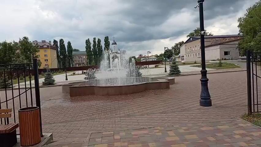 The video shows a fountain in a public square. The fountain has multiple jets of water spraying upwards and outwards, creating a misty effect. In the background, there is a small white building with a dome and a cross on top, likely a church or chapel. Tall green trees are also visible, along with some other buildings and a street lamp. The ground in front of the fountain is paved with brick-like stones. The sky is overcast with dark, heavy clouds, suggesting the possibility of rain. The scene is calm and serene.