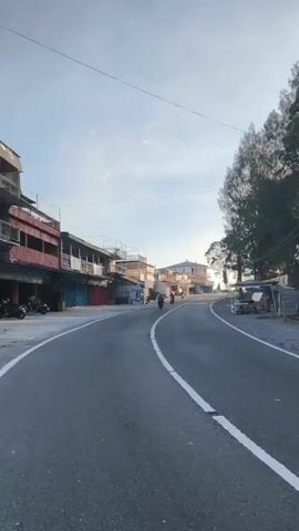 The video shows a road with buildings on the left and trees on the right. There are a few motorcycles on the road. The sky is clear with some clouds. The video appears to be taken from a moving vehicle. A sign that says "CEMOROSEWU" is visible in the foreground.