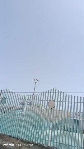 This video captures a forward-moving shot of a light blue, wavy fence with vertical slats. The fence is set against a backdrop of a clear, pale blue sky. Behind the fence, buildings with some red and white elements are visible, along with a tall lamppost topped with multiple lights. The fence appears to be made of metal and is mounted on a concrete base. There are two round, white circular signs attached to the fence. The scene suggests an outdoor setting, possibly a public area or along a road, given the movement of the camera. The time of day appears to be daytime, with bright sunlight. The weather seems fair, indicated by the clear sky.

Visible text includes a watermark at the bottom left corner reading "Infinix HOT 50 Pro". The concrete base of the fence also has some faint markings that are difficult to discern. The overall mood is neutral, focusing on the visual elements of the scene. The location cues, such as the style of fencing and the surrounding architecture, combined with the provided location context of Maiquetía, Venezuela, suggest a tropical or subtropical urban environment. No people are clearly identifiable in the video. The activity is simply the observation of the passing scenery.