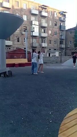 At 00:00, two young individuals are observed on a paved outdoor area in front of a multi-story building. One individual, wearing a white t-shirt and blue jeans, is actively engaged in an activity that appears to involve throwing or catching a dark-colored spherical object. Their body language suggests movement and exertion. The second individual, positioned slightly behind and to the right, is also facing the same direction and seems to be participating. A large, dark, cone-shaped structure on a pedestal is prominent in the left foreground. Behind the individuals, a section of the building is visible, characterized by its architecture and multiple balconies. The ground surface is a dark, textured material, likely asphalt or a similar composite. A wooden bench is also visible near the central cone structure. By 00:01, the dynamic of the scene has shifted slightly. The individual in the white t-shirt and jeans is now captured mid-movement, with their arm extended upwards and their body angled as if reaching for or throwing an object. The sphere they were previously interacting with is no longer clearly visible in the frame. The second individual is still in view, also looking in the same direction. The environment remains consistent, with the building and the large cone structure in the background and foreground, respectively. At 00:02, the perspective of the video has changed, providing a wider view of the outdoor space. The original two individuals are no longer the primary focus. Instead, the camera pans to reveal a group of people further in the distance. One person, wearing a white top and dark bottoms, stands near a low, curved structure. Other figures, including children, are scattered around the area. The background still includes the multi-story building, but also a distinct tree with dense foliage. The ground appears to be the same paved surface as before. By 00:03, the camera has continued its sweep of the area. The focus is now on a group of three individuals, appearing to be younger people, clustered together near the curved structure. One individual is wearing dark clothing, and another is in a light-colored outfit. They are interacting with each other, with one person holding what appears to be a rolled-up item, possibly clothing or fabric. The tree is prominent in the mid-ground, and more of the surrounding buildings are visible. A wire or cable is visible stretching across the upper portion of the frame. At
