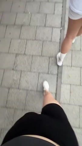 The video shows a person's legs and torso as they walk on a sidewalk paved with rectangular grey tiles. The person is wearing black cycling shorts and white sneakers. Another person's legs and feet, also wearing white sneakers, are visible further ahead on the right side of the frame. The camera angle is from a first-person perspective, looking down at the walker's legs and the ground. The overall impression is of two individuals walking side-by-side on a paved walkway.