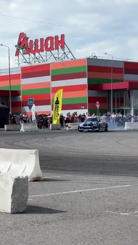 This media file captures a drift car event taking place in a parking lot outside a large retail building with a distinctive red and green striped facade. The building features a prominent "АШАН" (Auchan) logo at the top. The sky is partly cloudy, suggesting daytime. Spectators are gathered behind barriers, watching the action.

The primary subject is a blue drift car with intricate graphics and sponsor decals, including "diford" and "MIGHTY SHOW". The car is actively drifting, as evidenced by the smoke billowing from its tires. The event appears to be a competition or demonstration, with safety barriers and flags visible. In the background, a construction crane is visible, indicating potential ongoing development or construction in the area. The scene conveys excitement and the thrill of motorsport.