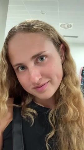 A close-up video shows a young woman with long blonde wavy hair and dimples. She is wearing a black tank top. Her eyes are closed and she has a slight smile. The background appears to be indoors with fluorescent lighting and a ceiling vent visible.