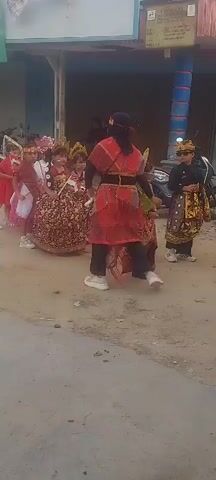 A group of children in traditional attire are participating in a parade. The children are dressed in colorful costumes, some in gold and red, with ornate headpieces. They are walking in a line down a dirt road, with adults in similar traditional clothing guiding them. In the background, there are buildings, motorcycles, and other spectators. The overall atmosphere appears to be festive, possibly for a cultural event or celebration.
