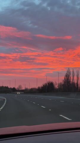 The video shows a view from inside a car driving on a multi-lane highway. The focus is on the sky at sunset or sunrise, which is a vivid mix of red, pink, and orange hues with some darker gray clouds towards the top of the frame. The road is asphalt with white lane markings and roadside lighting. Trees and shrubbery are visible alongside the highway. At least one other car can be seen driving in the opposite direction.