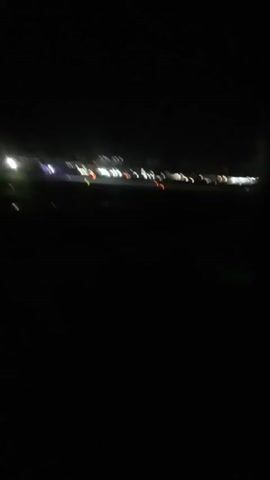 At night, from an airplane window, an airfield is visible, as well as lights on the plane's wing and on the ground. The lights on the ground appear to be from buildings and other vehicles. The scene is dark, with limited visibility due to the nighttime conditions. The plane seems to be moving, possibly taxiing or preparing for takeoff.