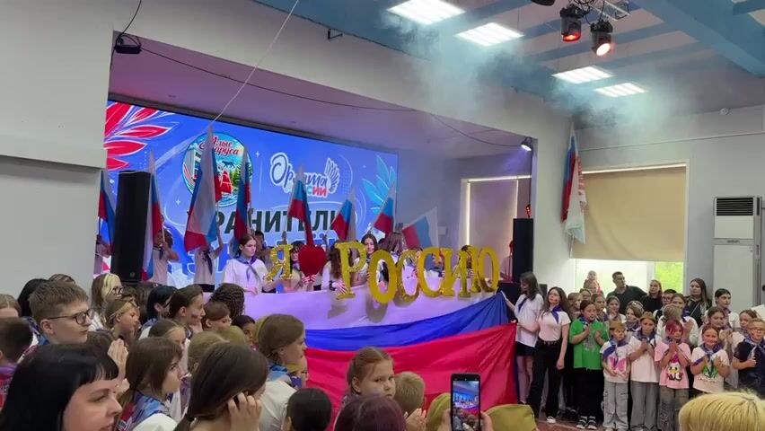 The video shows a group of young people gathered indoors, seemingly at an event or performance. A large screen displays text and graphics, including what appears to be a logo with the words "Орлята России" (Young Eagles of Russia) and "Хранители" (Guardians). In front of the screen, a group of individuals, primarily young women, stand behind a large banner that has the Russian flag colors. They are holding up large gold balloons that spell out "Я Россия" (I am Russia) and small Russian flags. The audience consists of many children and young adults, some of whom are wearing blue and white scarves, and appear to be clapping. There is some smoke or fog effects in the air near the ceiling lights. The environment looks like a hall or auditorium with stage lighting.