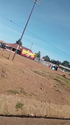 The video shows a truck parked in a dirt lot near a small town. The truck is white with a gray bed and has the words "ROOM BOOM" written on the side. In the background, there are several small buildings, some of which are painted in bright colors. There is also a power pole with a sign on it. The sky is blue and the weather appears to be sunny. The camera pans slightly to the right.