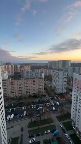 The video presents a panoramic view of a residential city area at sunset, likely taken from a high-rise building or balcony. The sky transitions from light blue at the top to warm orange and yellow hues on the horizon, with scattered clouds. In the foreground and midground, numerous multi-story apartment buildings, varying in color and style (some with pink and white facades, others in shades of grey and beige), dominate the landscape. Many cars are parked along streets and in parking lots between the buildings. Leafless trees line some of the roads. The overall impression is that of a calm, late afternoon or early evening in a densely populated urban environment. The specific location is Gavrilkovo, Russia. No people or discernible text are visible.