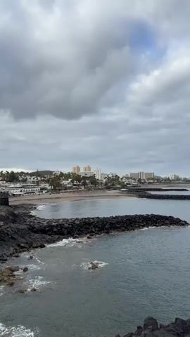 The video shows a coastline with a calm ocean under a cloudy sky. In the foreground, there are dark rocks jutting out into the water. In the middle ground, there is a beach with a few buildings and palm trees behind it. Further out in the ocean, there are breakwaters made of dark rocks. The sky is filled with grey clouds, with some patches of lighter sky visible. The overall mood is serene and peaceful.