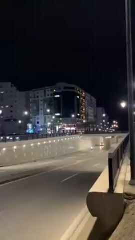 The video shows a nighttime urban scene. A road with a tunnel underneath the main street is visible. Cars and a scooter are driving on the road. Buildings are lit up in the background. A street light is on the right side of the frame.