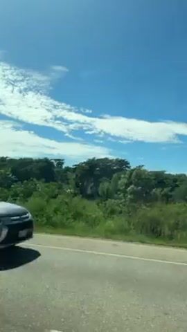 The video shows a vehicle driving down a paved road. The scenery features a bright blue sky with scattered white clouds and lush green vegetation. At one point, a white box truck passes the camera. The landscape is mostly flat, with occasional hills and trees.