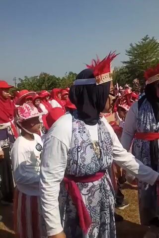The video shows a group of people, predominantly children and adults, participating in an outdoor event under a clear sky. Many individuals are wearing red and white attire, including headscarves, hats, and clothing with red and white stripes. Some participants, particularly adults, are adorned with elaborate headpieces featuring red feathers and gold accents. One adult woman in the foreground wears a black hijab, a white long-sleeved shirt, a patterned dress with a red sash, and sunglasses. She also has a large, dark accessory resembling a mustache attached to her face. Another adult woman next to her, also wearing a black hijab and a similar feathered headpiece, is looking and speaking towards the left side of the frame. A young child in the foreground, wearing a red and white outfit and head covering, is looking to the right. The background shows a larger crowd of similarly dressed individuals, suggesting a parade or a festive gathering. Some people are bending down, possibly preparing for an activity. The ground appears to be covered in dirt or grass, and trees are visible in the distance. The overall atmosphere seems to be celebratory.