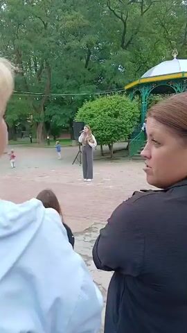 Two women are standing, facing away from the camera. The woman on the left has blonde hair and is wearing a light blue hooded jacket. The woman on the right has brown hair pulled back in a ponytail and is wearing a black jacket. They appear to be watching something happening in the background. In the background, there is an outdoor stage or gazebo with a group of people, some of whom appear to be children in uniform. A young woman is standing at a microphone on a stage, singing or speaking. There are trees and greenery in the background.
