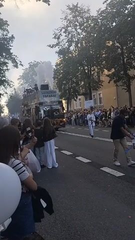 A slow-moving procession is depicted on a street lined with trees and buildings, with a large crowd gathered on both sides watching. The central focus is a large, open-top double-decker bus emitting a significant amount of white smoke from its upper deck. The bus has the words "LAUKINIAI VAKARAI" written on its side. People are visible on the upper deck of the bus, appearing to be participating in the event. On the street, a person in a white uniform stands facing the bus. The overall atmosphere suggests a parade or a festival. The video is shot from a low angle, with the camera positioned among the spectators.