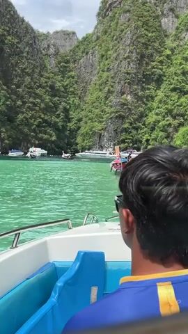 The video begins with a close-up view from the back of a white boat with blue seats, showing the upper back and head of a person with dark hair wearing sunglasses and a blue and yellow shirt. The boat is moving through a turquoise-colored body of water. In the distance, lush green, rocky cliffs rise steeply from the water, forming a scenic bay. Several other boats, including speedboats and longtail boats, are visible in the bay, some anchored and others moving. The person in the foreground is looking out towards the scenery. The camera then pans slightly to the left, offering a wider view of the bay, the cliffs, and the other boats. The water is calm, and the sky is overcast.