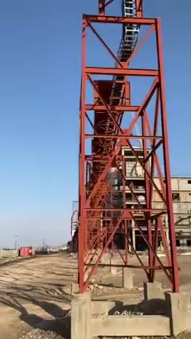 The camera pans upward along a red, steel structure towards a large, multi-story concrete building under construction.  The building has a tall, cylindrical structure on top. A crane is visible on the building. The ground around the building appears to be dirt and gravel. The sky is clear and blue.