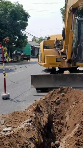 An excavator is digging a trench on the side of a road. The excavator is yellow and has a large bucket. The trench is filled with dirt and rocks. There are cars and houses in the background. A person is visible in the cab of the excavator.
