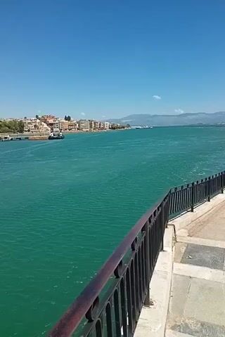 The video pans across a bright, sunny coastal scene. The foreground features a dark metal railing with a wooden top, bordering a paved walkway. Beyond the railing is a vast expanse of bright turquoise water, showing gentle ripples. In the distance, a coastline is visible with a row of white and tan buildings, some of which are several stories high. Trees and greenery are interspersed among the buildings. Further out, a narrow pier or jetty extends into the water, with a small boat docked or moored nearby. Mountains can be seen in the far distance, with a clear, pale blue sky overhead.