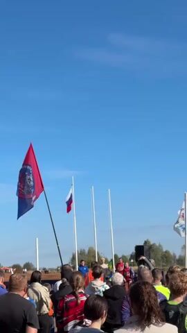 A group of people are gathered outdoors on a sunny day, with a clear blue sky above. Several flags are visible, including a red and blue flag with a crest, a Russian flag, and a white flag with blue and red elements. In the background, there are trees and what appears to be a stage or platform where several individuals, some wearing blue and red shirts, are standing. A speaker or microphone setup is present on the right side of the frame. The crowd is facing the individuals on the platform, suggesting an event or ceremony is taking place. Some individuals in the crowd are holding up their phones, likely to record the event. The overall atmosphere appears to be festive or celebratory.