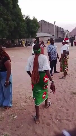 The video depicts a group of people in an outdoor setting. Most of them are standing and seem to be interacting with each other. The ground appears to be a dirt or sandy surface. A woman in a green skirt with red and white patterns, a white top, and a braided ponytail tied with a green bandana is a prominent figure, as the camera follows her. There are other individuals in the background, including some wearing white shirts and a person with a Santa hat. The environment includes trees and buildings, suggesting a village or community setting. The camera is moving, following the woman as she walks. The overall tone seems casual, as people are standing and talking.