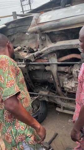 Two men in patterned shirts stand near an overturned vehicle. The vehicle is heavily rusted and appears to be undergoing repairs.  A tire rests on a part of the undercarriage. One man is touching his forehead. The location is outdoors, possibly a repair shop or garage. There are trees overhead.  Various tools and components are visible around the vehicle.