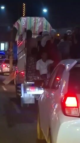 The video shows a group of people riding in the back of a truck at night. The truck appears to be a flatbed with a cage-like structure at the back, covered with a striped tarp. Several individuals are visible, sitting on the edge of the truck bed or standing inside. Some are wearing casual clothing, while others have head coverings. The truck is surrounded by other vehicles, suggesting it is in traffic or on a road. The lighting is dim, with red and blue lights from what appears to be a police vehicle visible in the background. The overall scene is crowded and appears to be a mode of transport for a large group of people.