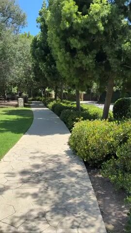 The video pans across a paved pathway in a park-like setting, likely in Beverly Hills, United States. The pathway is made of large, irregular stone pavers and curves gently to the right. On the left, a neatly manicured green lawn stretches out, bordered by a low stone wall and a metal fence. Beyond the lawn, trees and some covered seating areas are visible, suggesting a recreational space.

To the right of the pathway, a line of lush, green bushes creates a vibrant border, separating the path from the street or another area beyond. Behind the bushes, tall, leafy trees with dense foliage dominate the scene, casting dappled shadows on the path. The trees suggest a mature and well-maintained landscape.

The sky is a clear, bright blue, indicating a sunny day with fair weather. The lighting suggests it is midday or early afternoon, with the sun high in the sky. There are no people visible in the footage, giving a sense of tranquility and quietness. The overall impression is one of a peaceful, serene, and beautifully landscaped outdoor environment, perfect for a leisurely stroll. The video captures the natural beauty and manicured appearance of the location, characteristic of upscale residential or public areas.