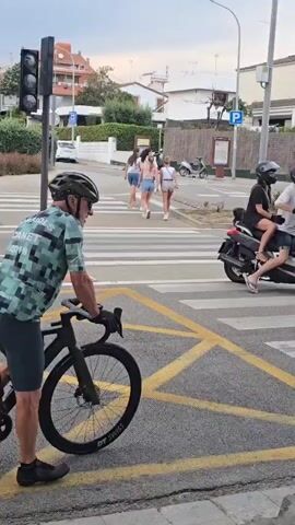 The video shows a crosswalk on a city street. Initially, a cyclist in a green patterned shirt and helmet is visible on the left side of the frame, waiting. A group of four pedestrians cross the street. A dark-colored scooter is also present, ridden by a person wearing a helmet. As the video progresses, the light turns red, and the scooter moves forward, carrying two people. One is wearing a white shirt and flip-flops. Several white cars, including a Citroen C3, are seen driving on the road in the background. The overall environment appears to be a residential area with various houses and trees visible in the backdrop. A parking sign is mounted on a pole near the crosswalk.