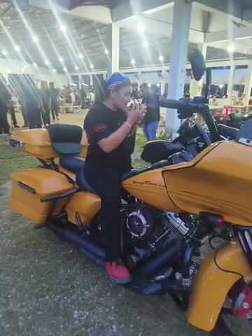A woman is sitting on a yellow Harley Davidson motorcycle. She is wearing a black t-shirt, black pants, a blue bandana, and pink shoes. She is wearing sunglasses and smiling at the camera. She raises her hand and flashes a peace sign and then a thumbs-up. The motorcycle is parked on grass under a covered pavilion. There are other people in the background, some of whom appear to be standing or sitting at tables. The lighting suggests it is evening or dusk.