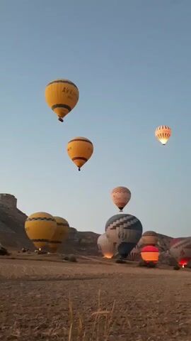 A video shows several hot air balloons in the sky at sunrise. Some balloons are in the air, while others are on the ground. The balloons are colorful and have various designs. The landscape below is rocky and arid. The sky is clear and blue with a hint of orange from the rising sun. The scene is peaceful and serene.