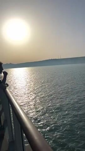 A serene seascape is captured from the deck of a vessel. The sun hangs brightly in the sky, casting a shimmering path across the water's surface. In the foreground, a dark brown railing runs along the edge of the deck, with a person's arm and hand visible resting on it. The distant shoreline is dimly lit, with faint silhouettes of structures and possibly smokestacks visible against the horizon. The overall atmosphere is peaceful, with the gentle movement of the water and the warm glow of the sun.