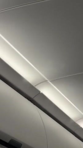 The video shows the overhead area inside of what appears to be a commercial airplane. The camera is positioned looking upward at the overhead compartments and ceiling, which are white with recessed lighting. A person with short blond hair walks past the camera briefly.  Yellow warning stickers are visible on the side of the overhead compartment.