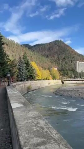 The video showcases a serene scene of a river flowing next to a paved walkway with a low concrete wall, with lush trees and a mountain in the background under a partly cloudy blue sky. The river water is moving rapidly and appears slightly rocky. The trees include evergreen pines and deciduous trees with some yellow leaves, suggesting it's autumn. A suspension bridge is visible in the distance. A tall building stands adjacent to the mountain. There are lamp posts and benches along the walkway, indicating a park or recreational area. The camera moves slightly to the left during the video.