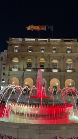 This is a vertical, nighttime video of a public square with a fountain and a building. The fountain in the foreground is illuminated with red and green lights, with water spraying upwards in various patterns. Behind the fountain is a stone building with arched walkways on the ground floor and windows on the upper floors. At the top of the building, there are several flags flying, including the Spanish flag. The sky is dark, indicating it is nighttime. There appear to be a few people in the background, but they are not clearly visible. The video is stable and appears to be filmed from a stationary position.