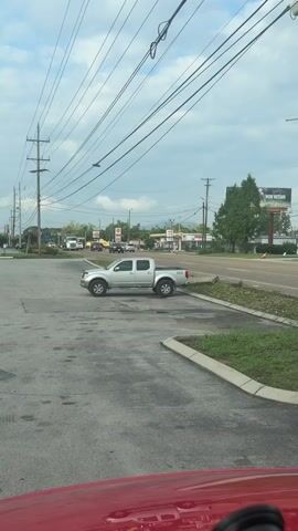 A silver Nissan Frontier pickup truck is parked in a parking lot. Power lines are visible overhead, as are telephone poles. In the background, there is a road with cars and trucks. A gas station is also visible in the background, as is a billboard. The sky is cloudy. The video is shot from inside a red vehicle, as evidenced by the red hood and dashboard visible in the foreground.
