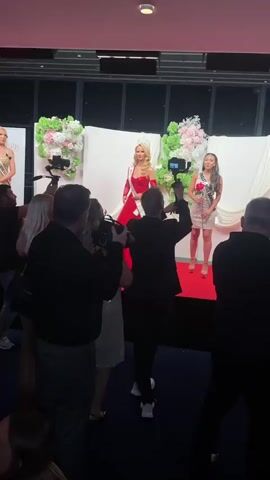 The video shows a beauty pageant stage at what appears to be an indoor event. Three women wearing pageant sashes, crowns, and roses are standing on a red carpeted stage in front of white backdrop with floral decorations. A crowd of people stands in front of the stage, and several photographers and videographers are actively capturing the scene. The camera moves slightly, capturing different angles of the event. The lighting in the room is subdued with a reddish hue, likely from stage lighting.