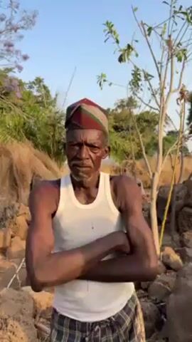 A medium shot shows an older, dark-skinned man wearing a white tank top and a patterned cap. He has a mustache and is staring at the camera with his arms crossed. The background features a dry landscape with scattered rocks, sparse vegetation, and a bright, slightly overcast sky. The setting suggests a rural or less developed area.