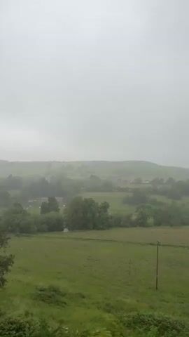 The video shows a view of a green, rolling landscape under a hazy, overcast sky. In the foreground, a grassy field is visible with a single wooden post standing vertically in the center. Further back, scattered trees and foliage create a textured layer. In the middle ground, the landscape continues with more trees and fields, and hints of buildings or structures are visible in the distance, nestled within the hills. The overall impression is of a rural, possibly damp, day. The camera appears to be moving, as evidenced by the slight shift in perspective and the blurring of trees in some frames.