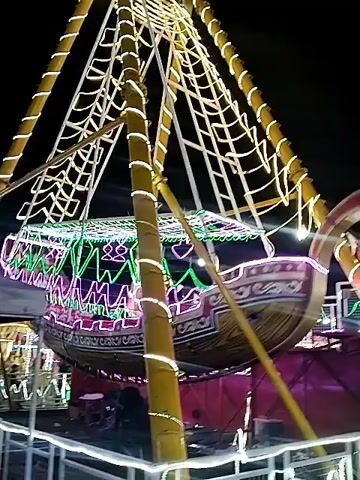 Here is a description of the video. 

The video shows a pirate ship amusement ride at night. The ride is decorated with bright neon lights in green, purple, and white.  The camera angle is low, looking up at the ride as it swings. The ride appears to be made of bamboo or a similar material. There are people on the ride.  A white fence surrounds the base of the ride.  In the background, there is a small tent or canopy with people standing underneath it. The sky is dark.