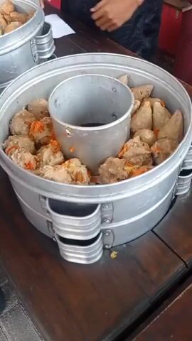 A close-up shot focuses on a large, multi-tiered aluminum steamer filled with meatballs. The meatballs vary in size and shape, with some being round and others more conical. They are arranged around a central cylindrical insert within the steamer. Scattered amongst the meatballs are pieces of what appear to be chopped chili peppers, adding a vibrant orange-red contrast. The steamer itself is placed on a dark wooden surface, possibly a table. In the background, a person's arm and shoulder are visible, suggesting someone is operating or attending to the steamer. Another similar steamer, also filled with meatballs, is partially visible in the upper left corner of the frame. The overall impression is of a food preparation or serving station.