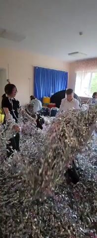 The video shows a room filled with silver tinsel streamers. There are multiple people in the room, including children and at least one adult. The children are throwing handfuls of the tinsel. The adult is also handling the tinsel. There are some furniture items in the room, including yellow beanbag chairs, a black umbrella, and possibly other items obscured by the streamers. The room is lit by overhead fluorescent lights. There is a blue curtain partially covering a window. Overall, the scene appears to be festive and celebratory, albeit somewhat messy.