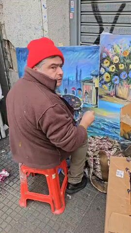 Here is a concise description of the video:

The video shows a man painting on the street. He is sitting on a red plastic stool. He is wearing a brown jacket, khaki pants, black shoes, and a red beanie. He is holding a paint palette in his left hand and a paintbrush in his right hand. In front of him are two canvases. The one on the left depicts a landscape with a train, a building, and people with umbrellas. The one on the right is a painting of flowers in a vase. Behind him is a metal shuttered door and a cardboard box. The ground is tiled.