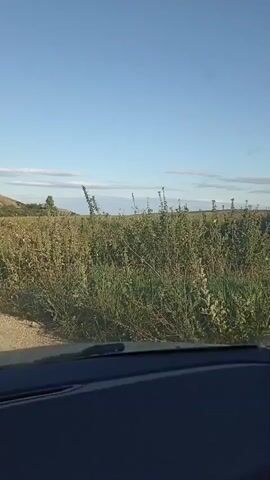 The video shows a view from inside a vehicle driving down a dirt road. The road is unpaved and appears to be in a rural area. On either side of the road, there are fields of tall grass and vegetation. In the distance, there is a large, rolling hill covered in greenery. The sky is clear and blue with a few wispy clouds. The camera is pointed forward, capturing the road ahead and the surrounding landscape. The vehicle is moving at a moderate pace, and the scenery is gradually changing as it progresses. The overall impression is of a peaceful, scenic drive through the countryside.