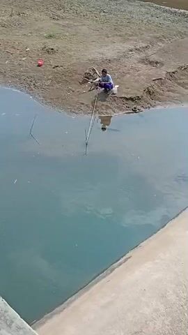 A person is fishing on the bank of a body of water. The person is crouching and holding a fishing rod that extends into the water. There is a white bucket and some debris near the person. The ground is muddy and appears to be a riverbank. The water is murky.