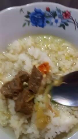 A close-up shot shows a bowl of rice porridge with meat and chili. A spoon is in the bowl, lifting some of the porridge. The bowl has a floral pattern.