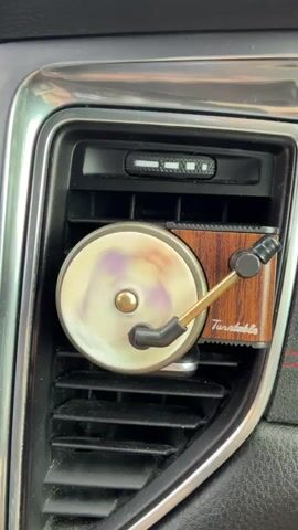 The video shows a close-up of a car's air vent. Attached to the vent is a novelty air freshener designed to look like a miniature record player. The record player's turntable is spinning, indicated by the blurred purple and white colors on its surface. The arm of the record player is extended, and the entire air freshener is attached to the car's vent. The surrounding environment appears to be the interior of a car, with the vent and surrounding dashboard elements visible.