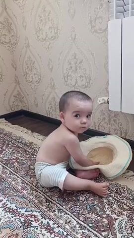 A baby is sitting on a rug in front of a wall and a radiator. The baby is wearing a diaper and is reaching into a straw hat. The baby looks into the hat and then looks at the camera. The baby then reaches into the hat again. The baby is in a room with a patterned wallpaper and a patterned rug.